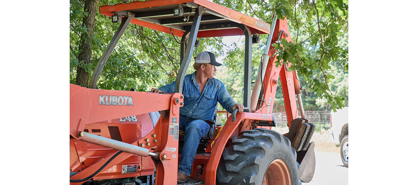 Un hombre, Wes Stevenson, con una camisa de mezclilla y una gorra, está sentado en un tractor naranja bajo árboles frondosos, mirando hacia un lado mientras opera el equipo.